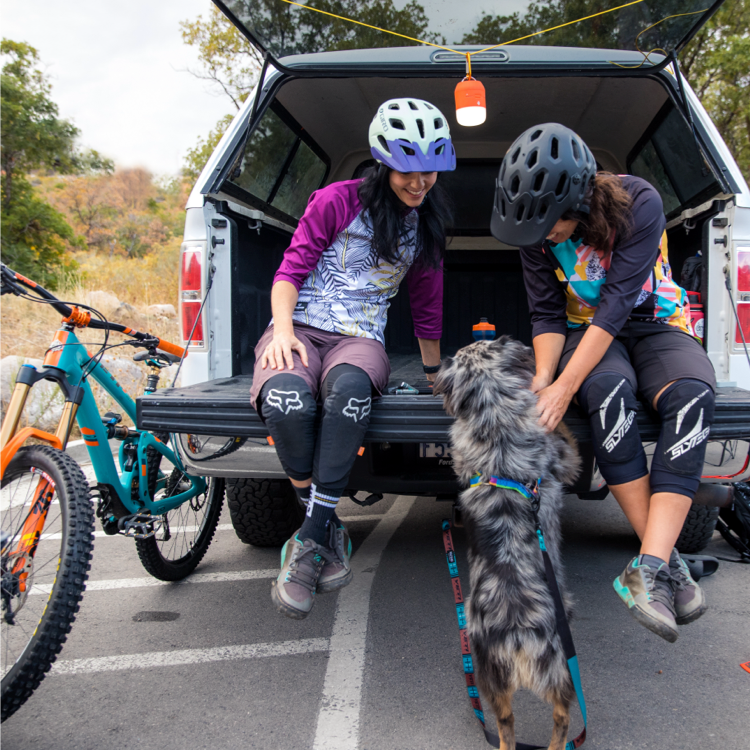 Two women mountain bikers sitting on a tailgate petting a dog