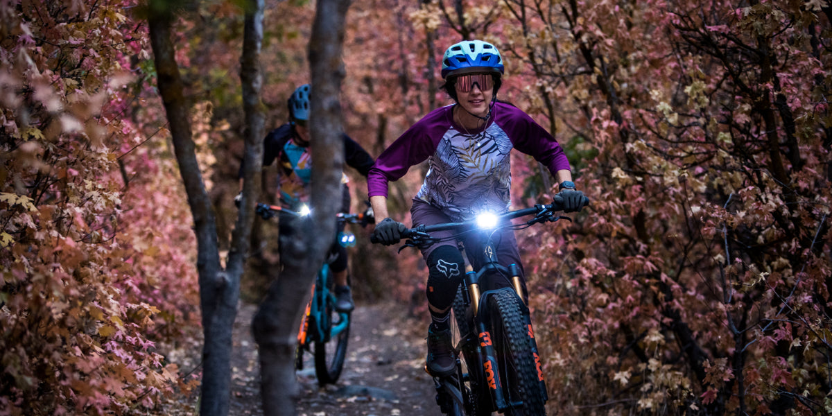 Two women mountain biking in a forest with bright headlights